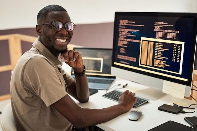 man with glasses looking into camera with computer screen showing some code samples in the background.